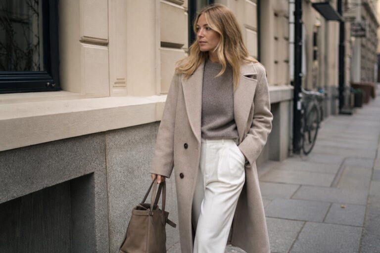 Scandinavian style fashion outfit with neutral coat, knit sweater, tailored trousers, and clean leather sneakers on a city street